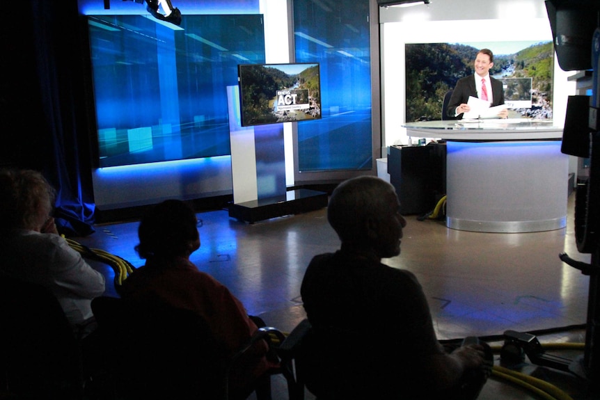 Wide shot of ABC News studio with newsreader smiling at Elders watching news go to air.