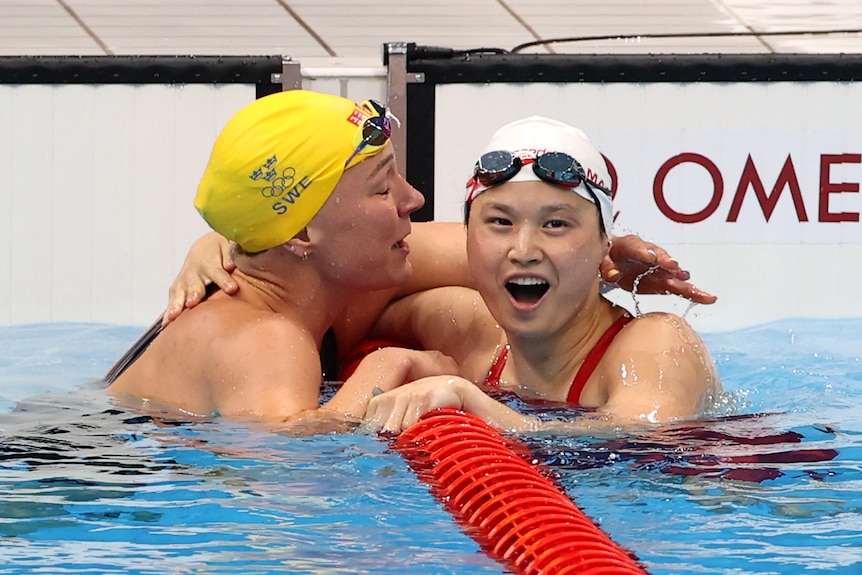 A woman wearing a white swimming cap is hugged by another woman wearing yellow swimming cap in a pool