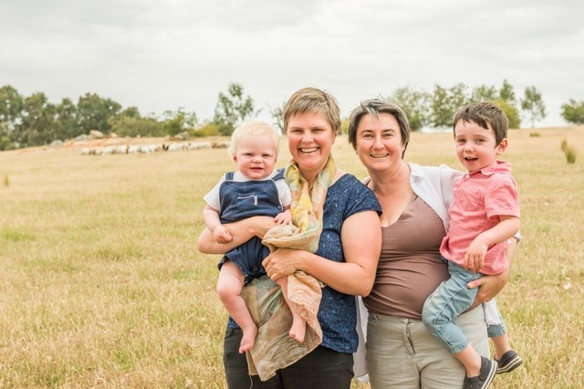 lady in blue top and scarf smiling holds baby in blue overalls next to lady in brown top holding child in open paddock