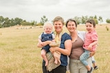 Lady in blue top and scarf smiling holds baby in blue overalls next to lady in brown top holding child in open paddock.