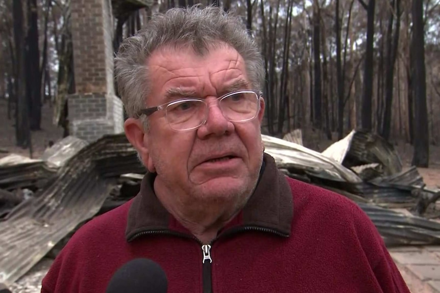 A man wearing glasses stands in front of the burnt ruins of his home.