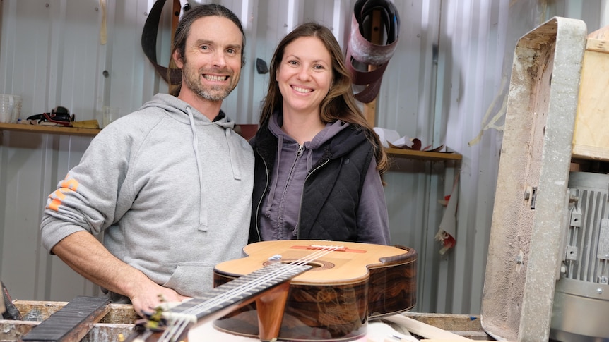 The pair stand in a workshop near a guitar