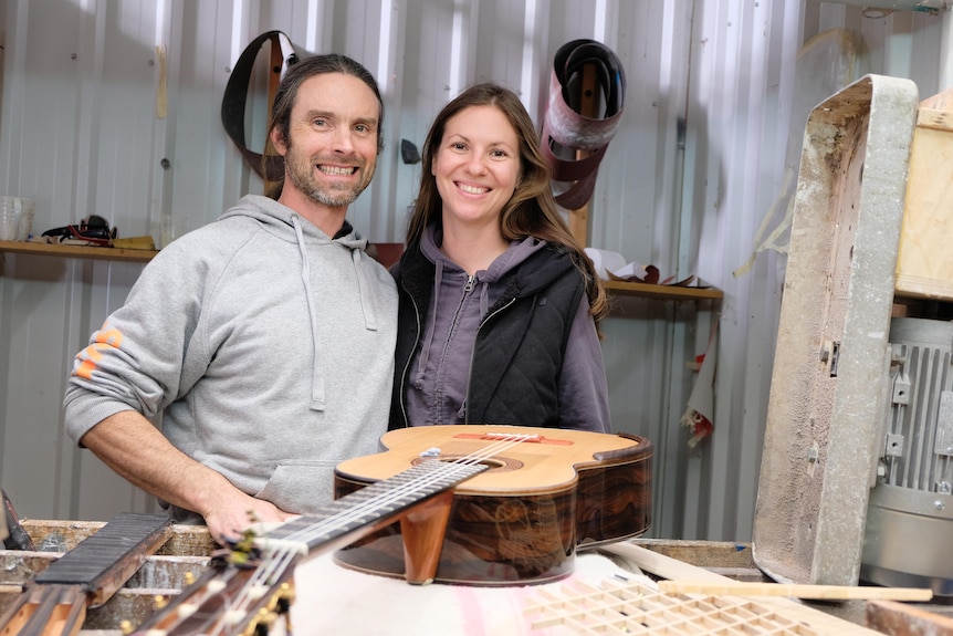 The pair stand in a workshop near a guitar
