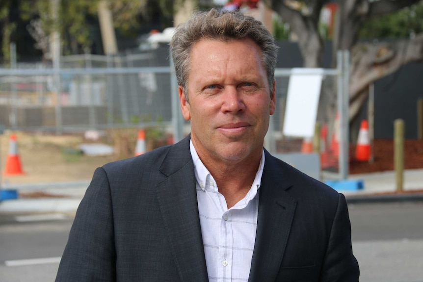 Gavin Hegney poses for a photo in front of a road wearing a grey suit jacket and a white shirt.