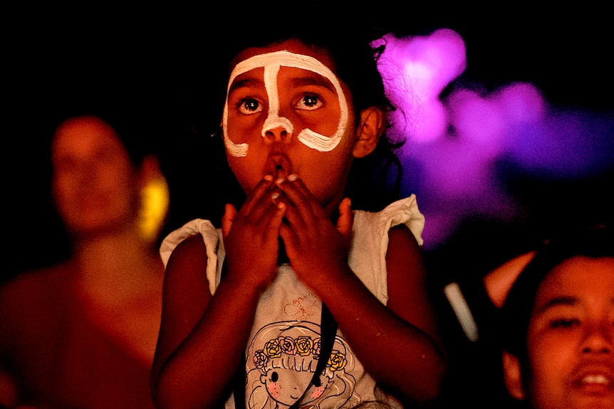 A young Indigenous girl sits on the shoulders of a woman, as they watch the live music at the NIMAs.