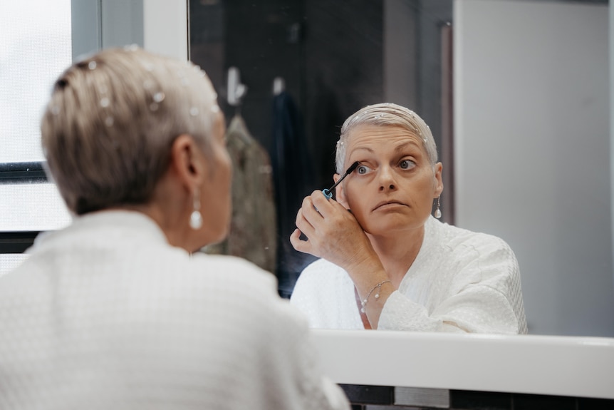 A woman in a white jacket looks in the mirror as she applies makeup.