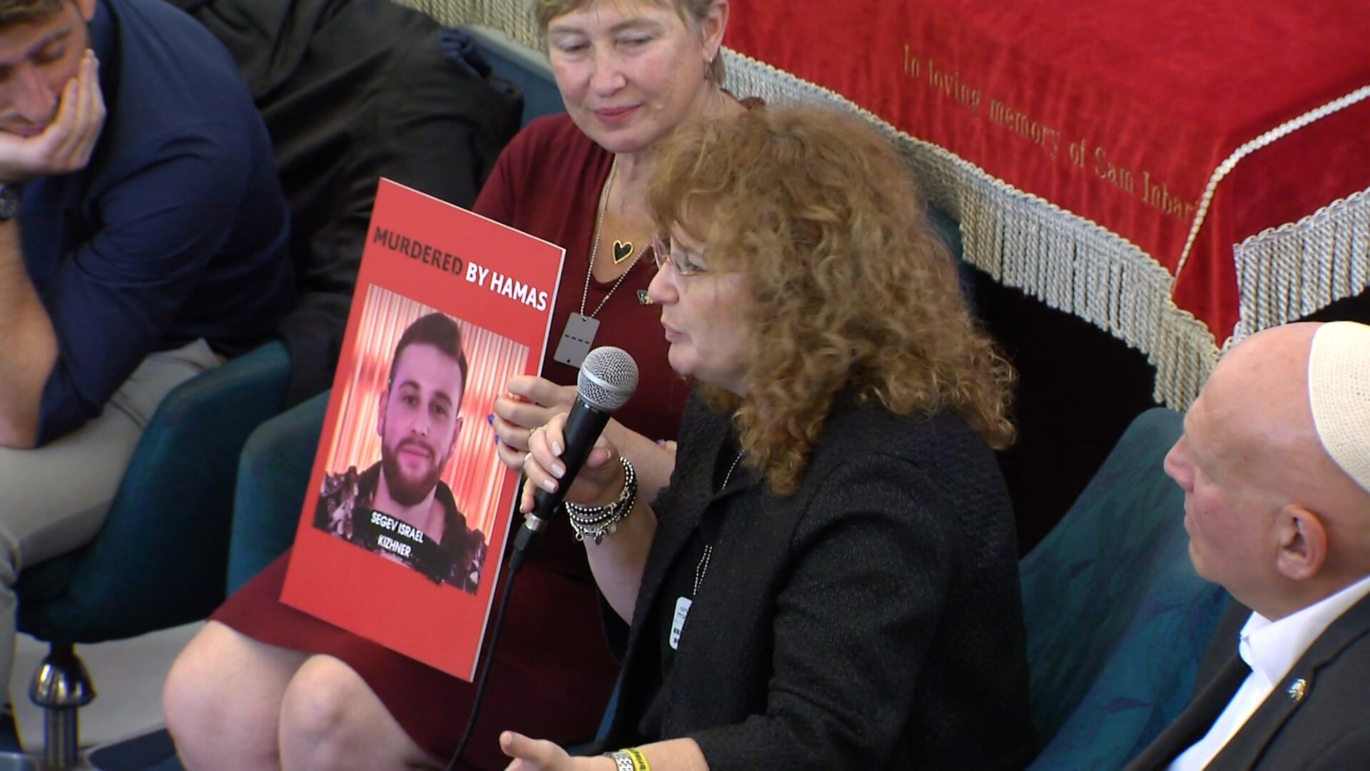Tali Kizhner, a woman with brown curly hair and glasses holding a microphone. A photo of her son next to her
