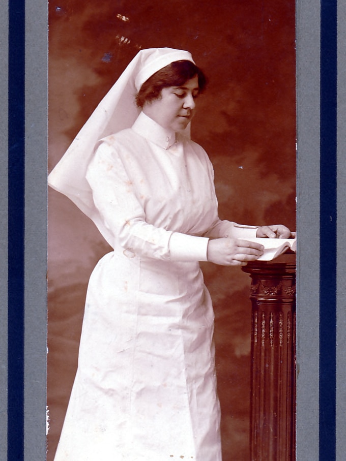 An historic photos of a woman posing reading a book in her nurses uniform