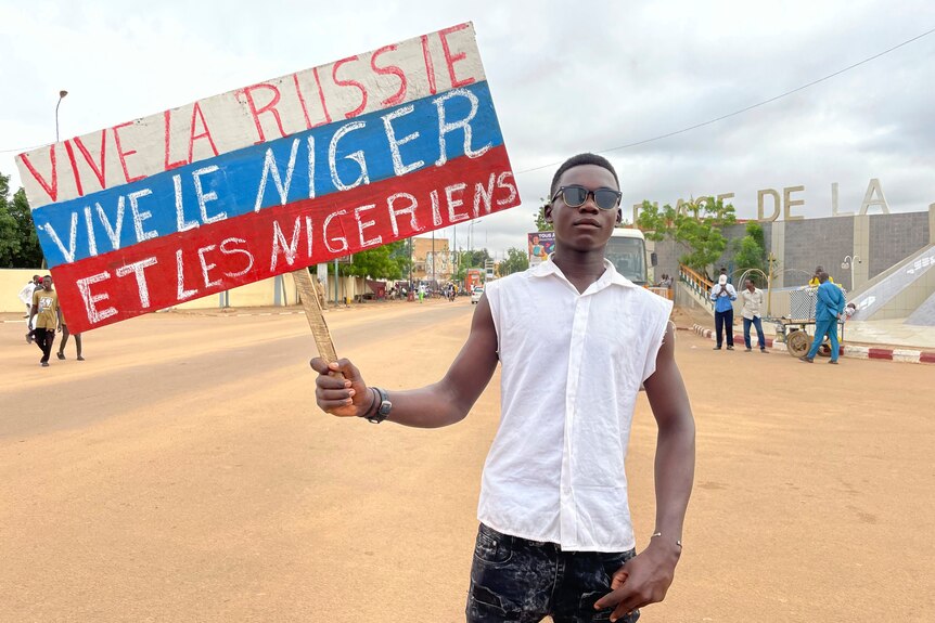 Man in white shirt and sunglasses looks serious while holding sign that says "Long Live Russia, Long Live Niger and Nigeriens".