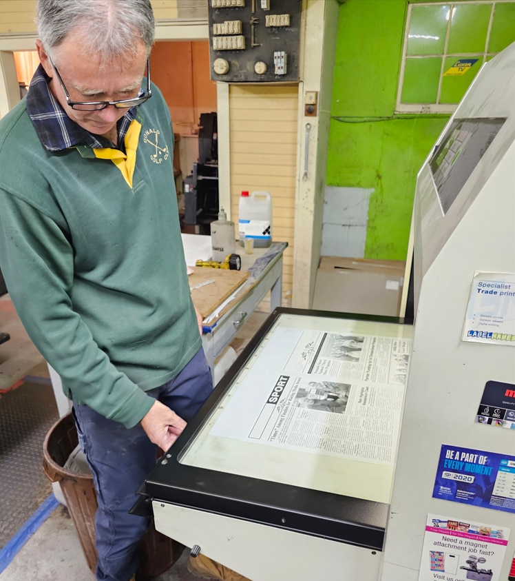 A man stands next to a printing press with a newspaper page on it, he looks down and presses a button.