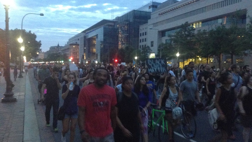 A large crowd of people walk up a street, many of them holding placards.