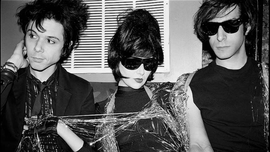 Black and white photo of Yeah Yeah Yeahs sitting on a couch, covered in shiny streamers. Karen O wears sunglasses and big hair.