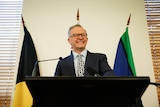 Anthony Albanese smiles while standing behind a podium with the aboriginal, australian and torres strait island flags behind him