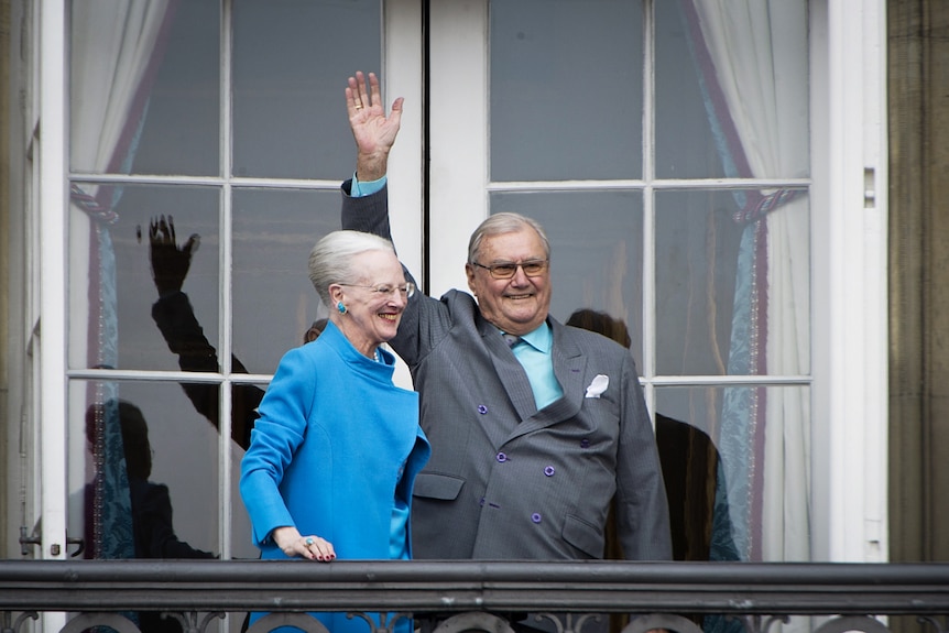 Queen Margrethe in blue coat and matching earrings smiles with Prince Henrik who waves from balcony