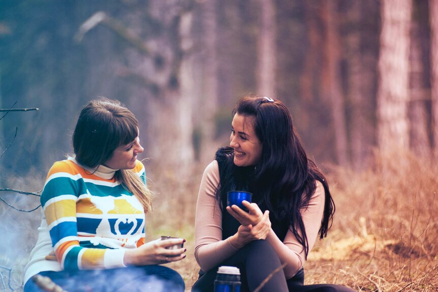 Two smiling women sitting on the ground chatting while camping in the bush.