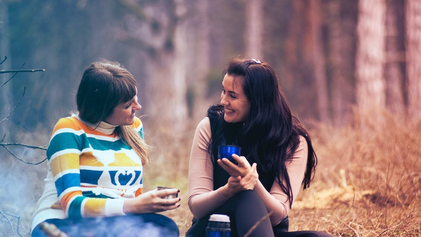 Two smiling women sitting on the ground chatting while camping in the bush for a story about eco camping tips.