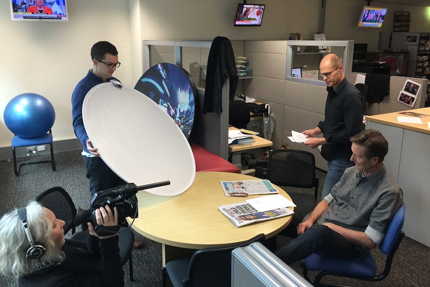 Jake Murphie holding bounce board as Julie Adams films Tim Latham talking to Paul Barry seated at table in office.