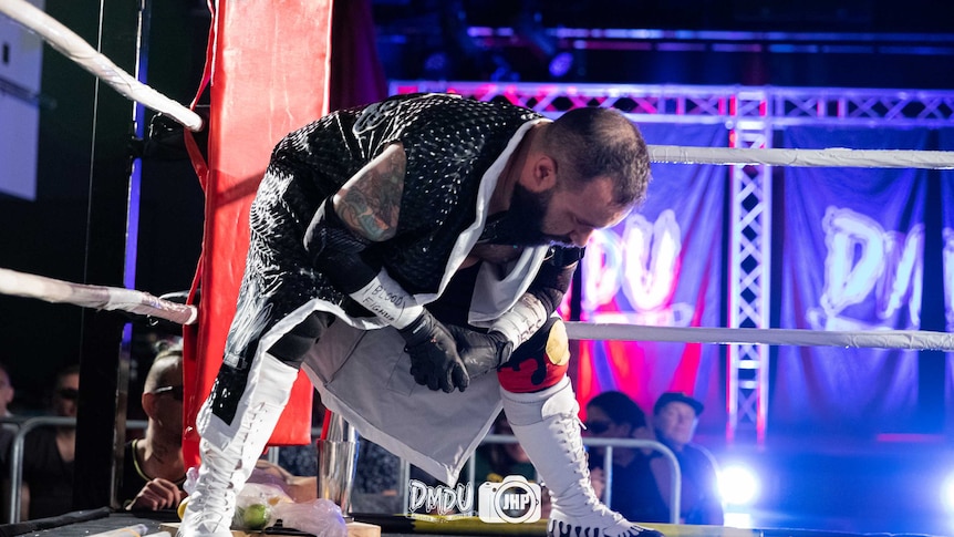 In a boxing ring, a man wearing a black cape with mid-calf white boots bows. The words 'blood fighter' are written on his arm.