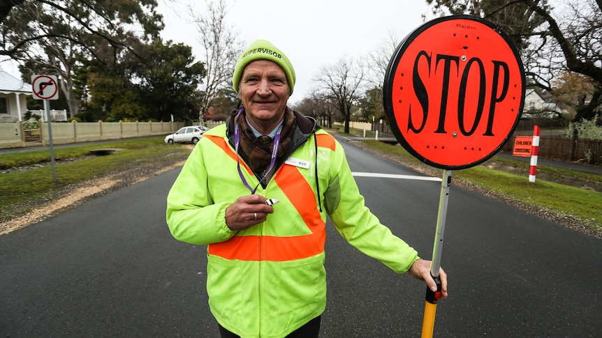 Crossing guard Ray Stevenson stands in middle of the road. He holds a stop sign in one hand, a whistle in the other.