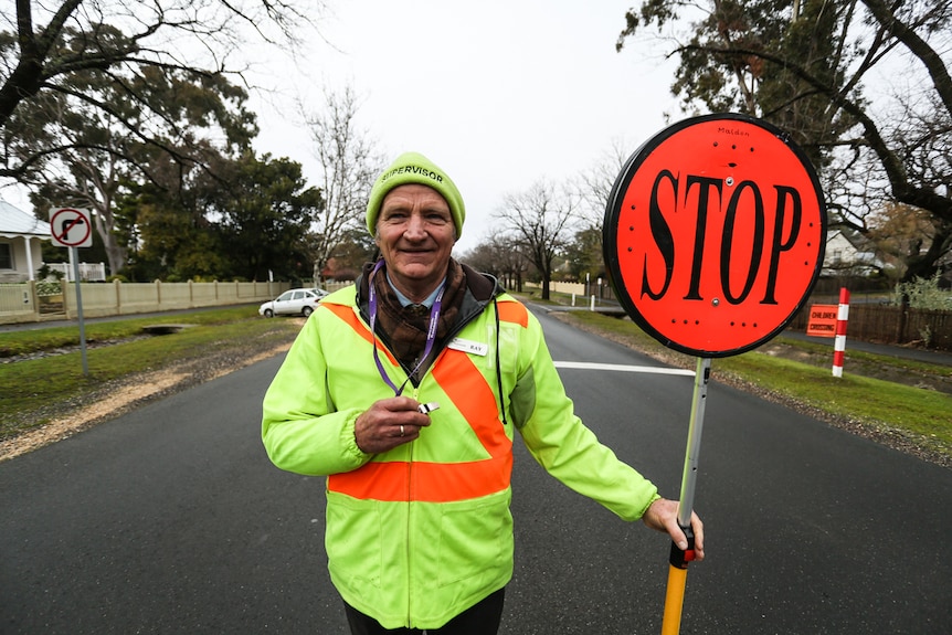 Crossing guard Ray Stevenson stands in middle of the road. He holds a stop sign in one hand, a whistle in the other.