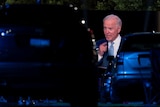 Cars parked infront of Joe Biden speaking on a screen at a baseball stadium