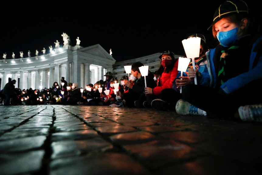 People sit outside on stone pavement holding candles, white building in background.