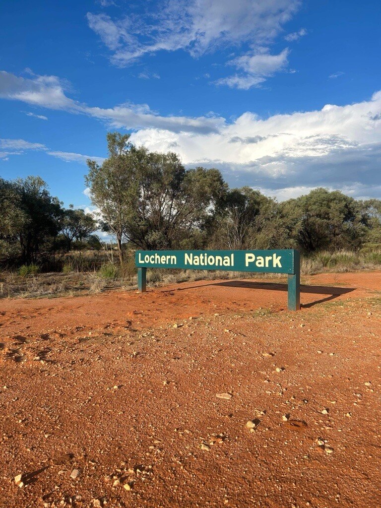 A green sign says Lochern National Park on red sand with trees and blue sky behind. 