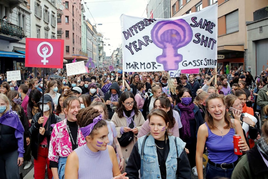 a crowd of women wearing purple items and holding signs walking down a street in Zurich