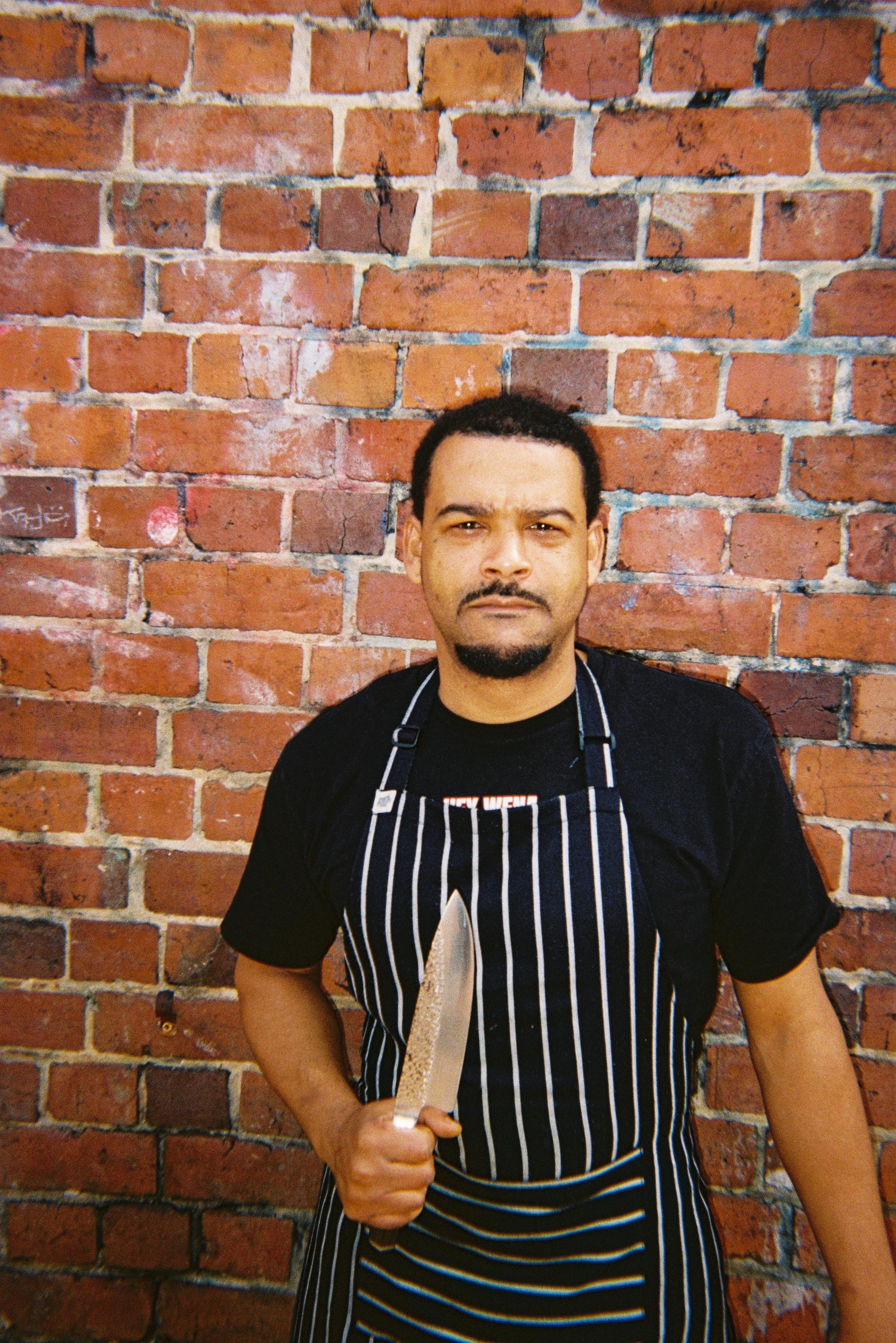 Un hombre con un delantal a rayas se para frente a una pared de ladrillos y sostiene un cuchillo de chef.