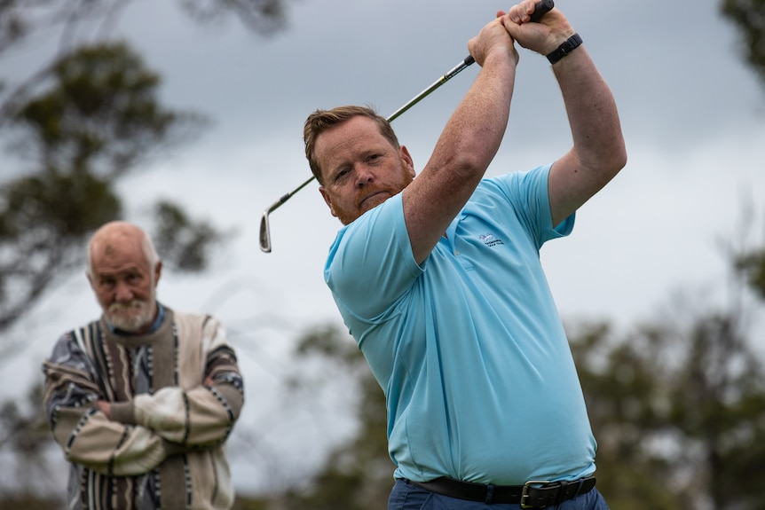 A man takes a shot with a golf club as another man watches from behind