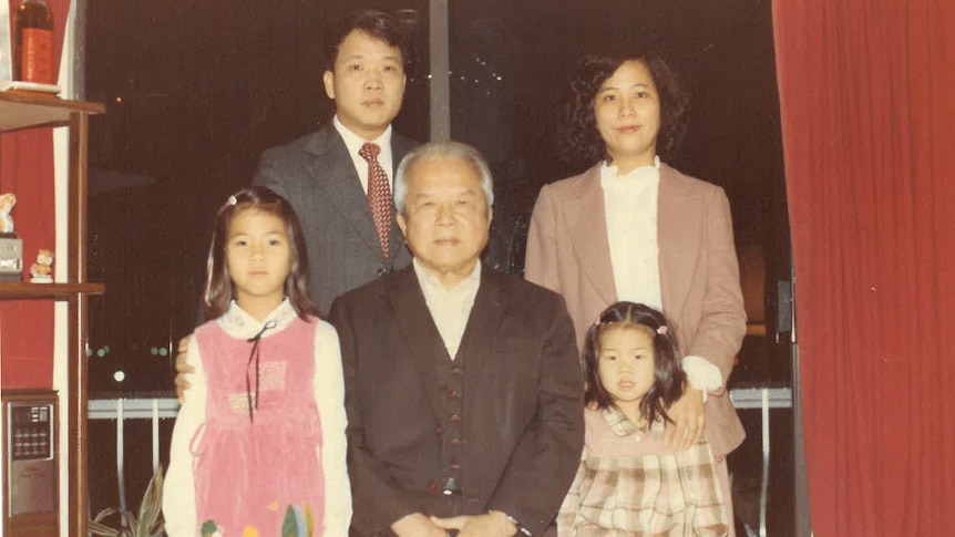 Slightly grainy family photo of two adults standing at back, two young girls in front and in the middle an older man. 