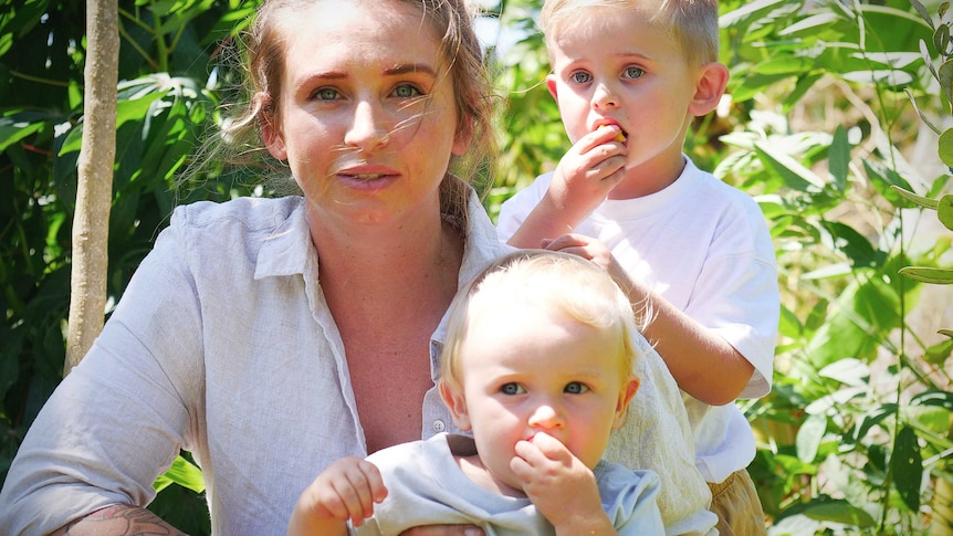 Mother sitting in garden and holding baby and toddler at her shoulder eating fruit, in a story about birth trauma.