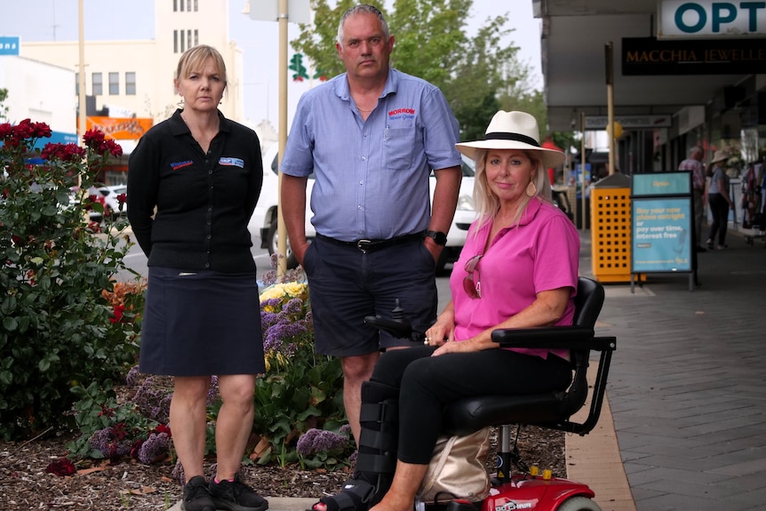 a woman with blonde hair, black jumper stands next to man with blue shirt, next to a woman in pink shirt on motorised scooter