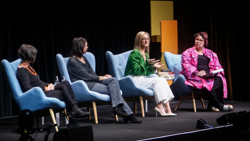 Four women in a row onstage in chairs, talking. Names in caption