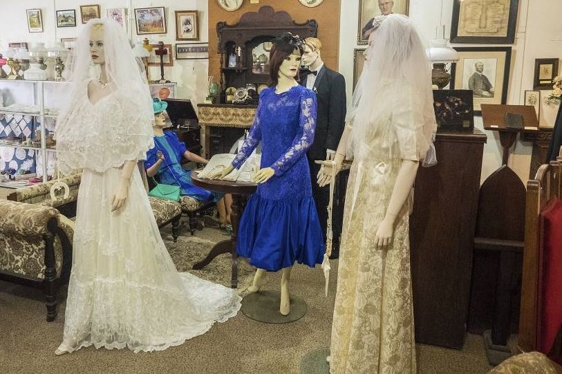 Several mannequins dressed in wedding gowns with other museum pieces in the background.