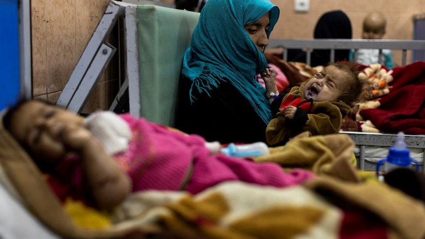 Small sick child wrapped in pink blanket lies on hospital bed in foreground beside woman in teal hijab nursing baby