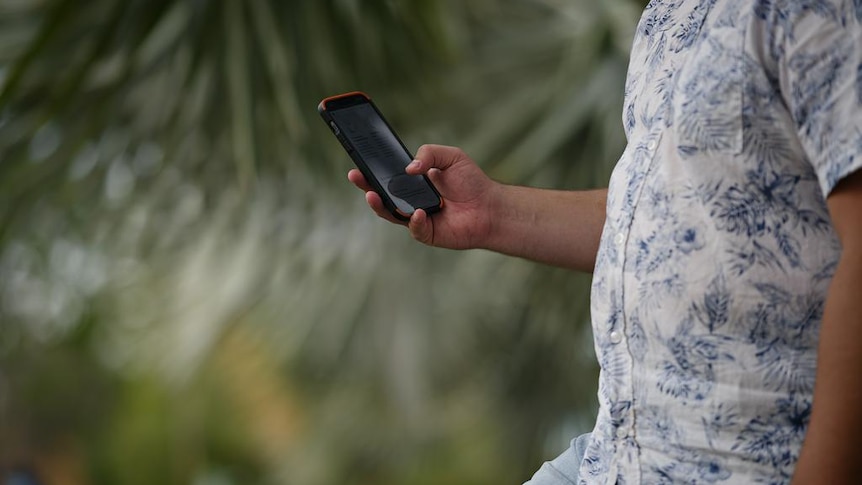 A man's hand holding a mobile phone. His torso is in shot and there are out-of-focus plants behind him.