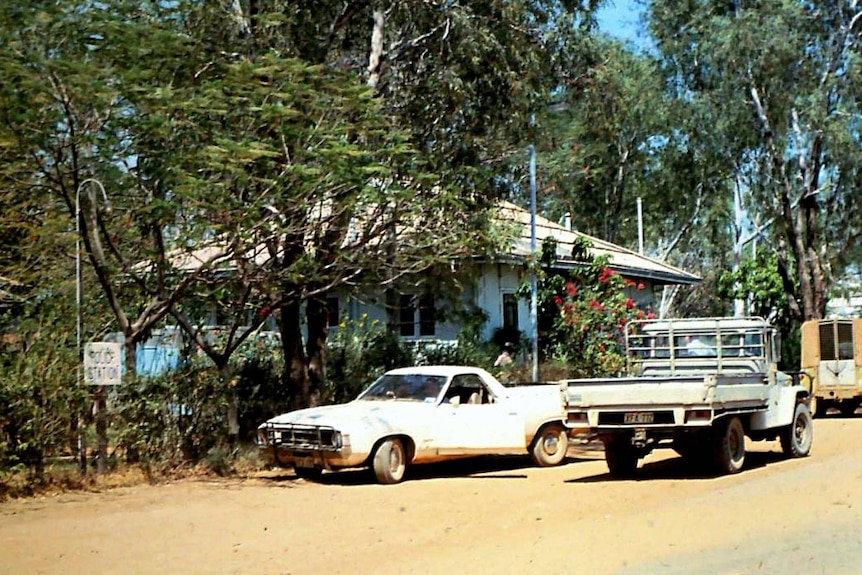 Two utes are parked on a dirt road outside a building.