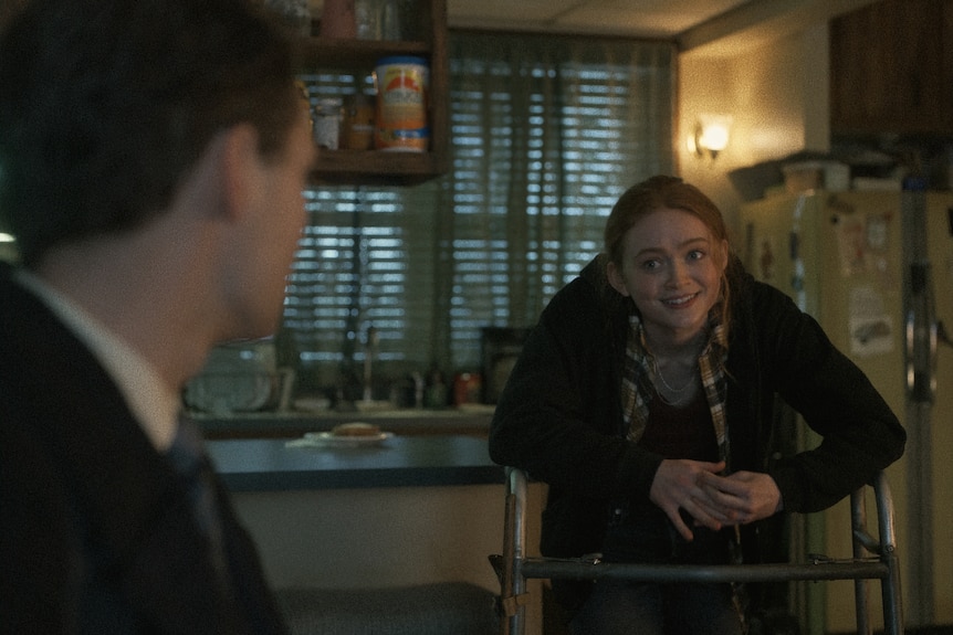 Young white red-haired girl leans on a zimmer frame in a dark kitchen while talking to a young white man in the foreground.