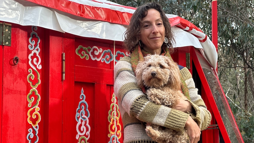 A woman with curly hair, carrying her dog, standing outside a colourfully-decorated red yurt.