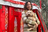 A woman with curly hair, carrying her dog, standing outside a colourfully-decorated red yurt.