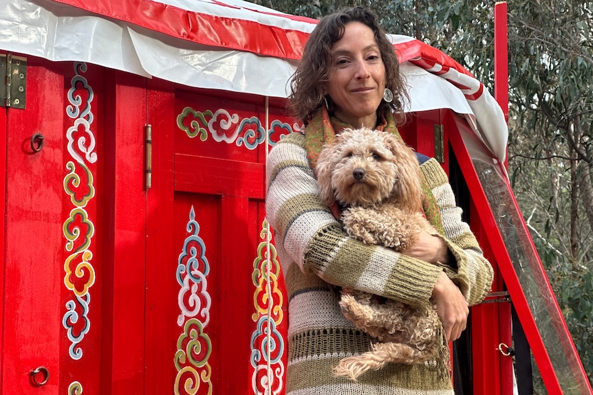 A person carrying her dog, standing outside a colourfully-decorated red yurt.