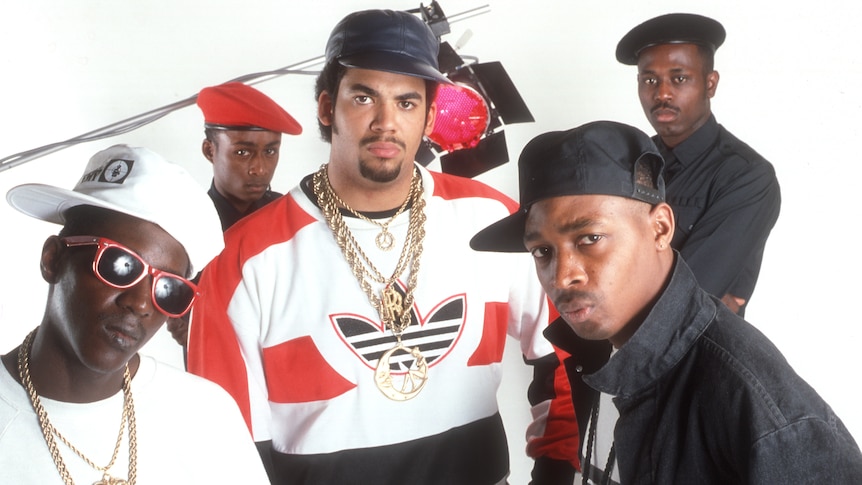 Five members of Public Enemy pose in a white photography studio looking up into camera
