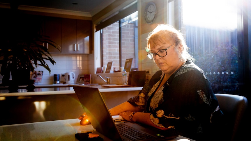 Rachel sitting at her kitchen table using her laptop. She is blonde and wears glasses.