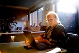 Rachel sitting at her kitchen table using her laptop. She is blonde and wears glasses.