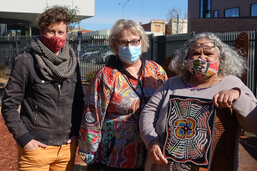 Three people wearing masks stand outside on a sunny day.