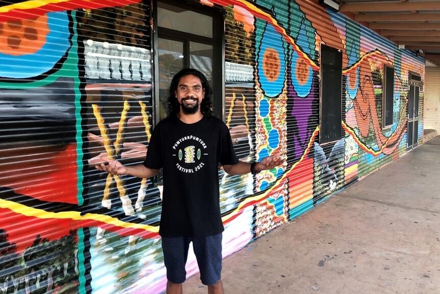 A smiling man with long hair and a beard, standing in front of a mural.