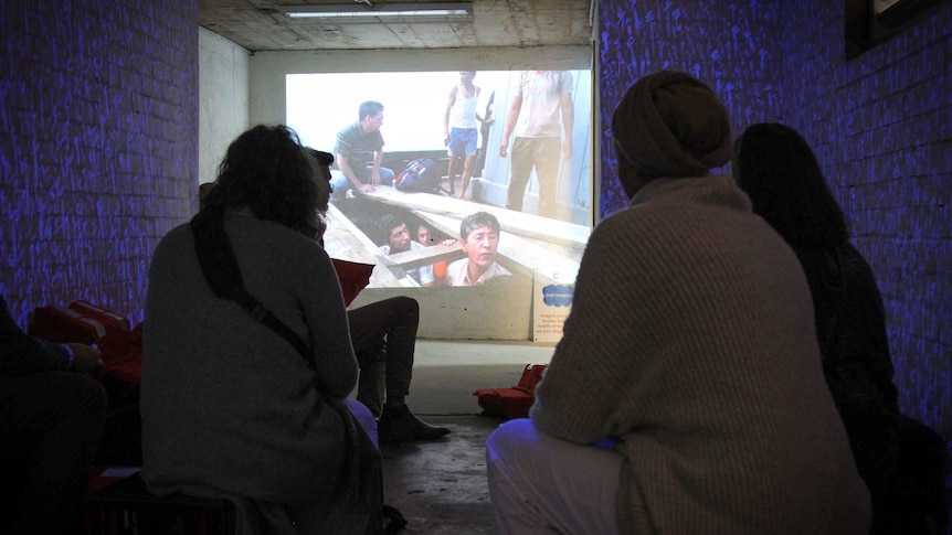 A basement with blue lights used to make it look like water, and a screen with a photograph of a people smugglers boat.