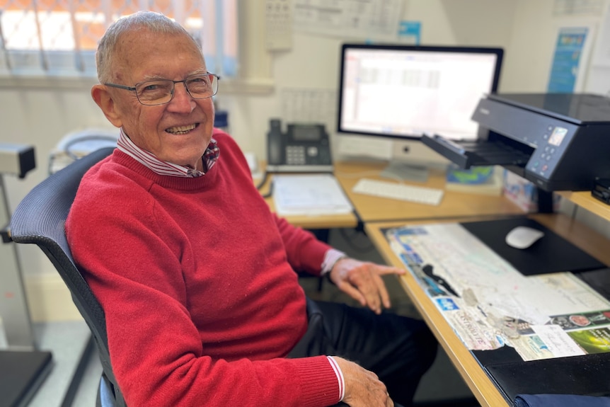 An older doctor in a red jumper sitting at a desk.