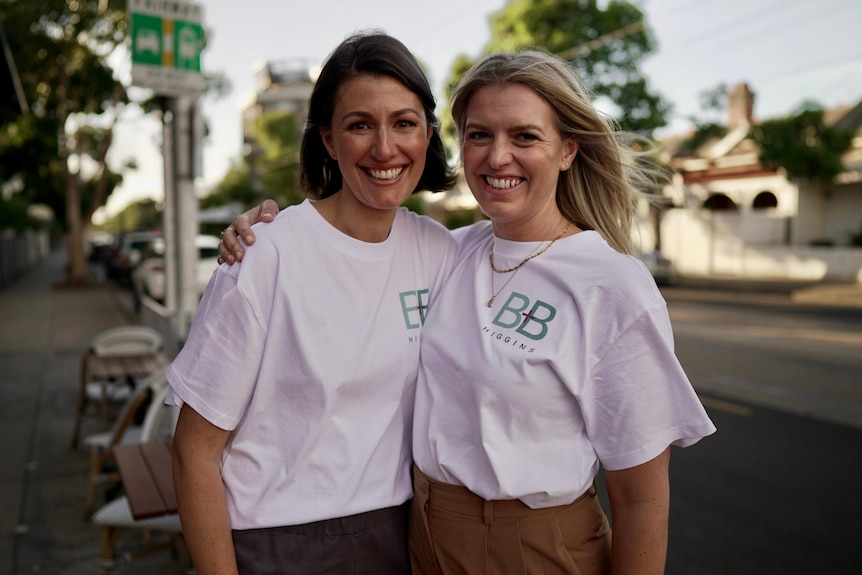 Two white women wearing white political campaign t shirts stand and smile by the side of a road